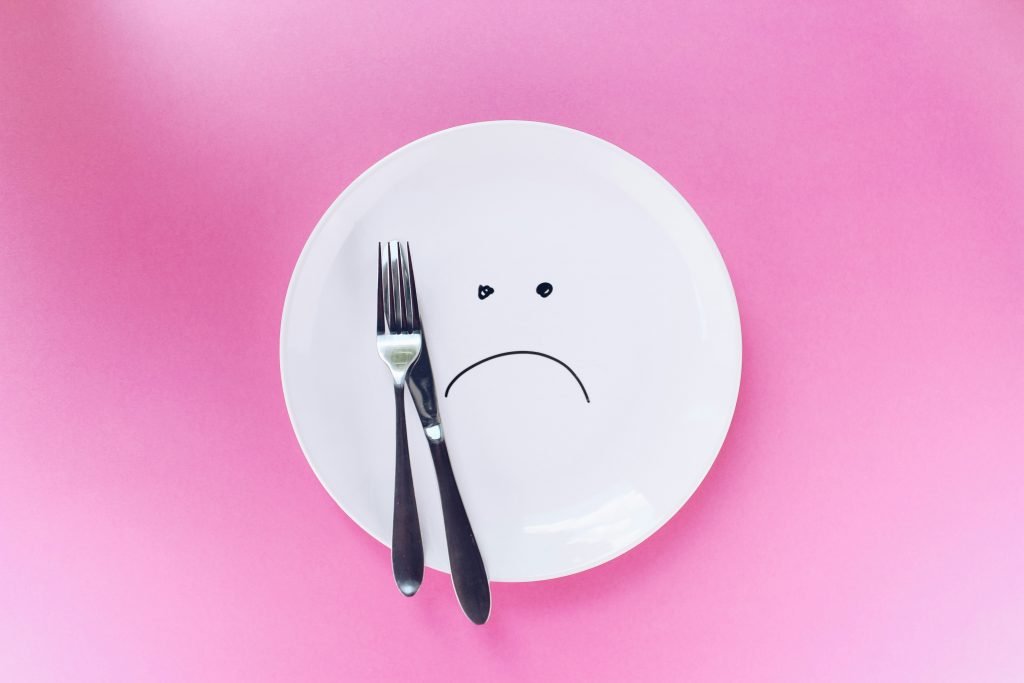 Ein leerer Teller auf pinkem Hintergrund zeigt ein trauriges Gesicht aus Besteck und aufgemaltem Mund. Stimmungsschwankungen und Appetitlosigkeit sind häufige Begleiterscheinungen bei ADHS Schweiz und beeinflussen oft die Essgewohnheiten.