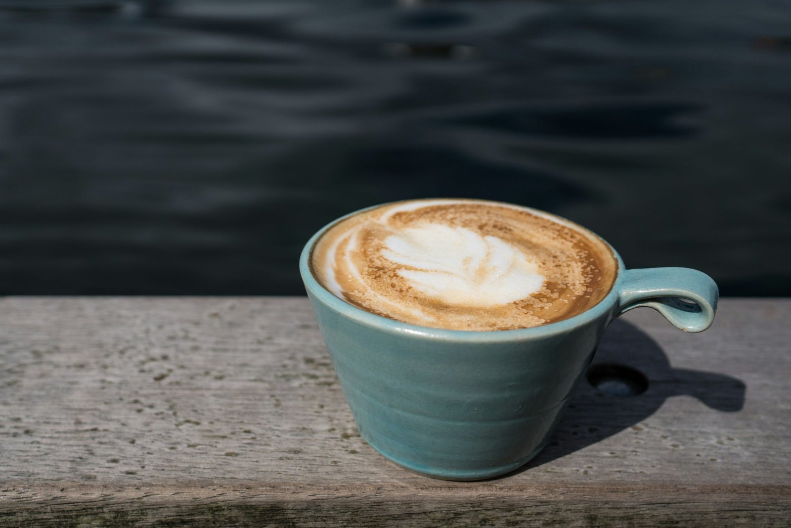 Eine Tasse Cappuccino mit schöner Latte-Art steht auf einer Holzbank am Wasser. Das ruhige Ambiente symbolisiert eine entspannte Pause im Alltag. Auch Menschen mit ADHS Schweiz können von bewussten Ruhepausen profitieren.